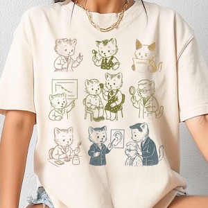 Op de afbeelding: Cr&egrave;mekleurig T-shirt met een herhaald patroon van cartoonkatten in verschillende doktersposes. De katten zijn afgebeeld in een vintage stijl, dragen laboratoriumjassen en gebruiken medische instrumenten. Het ontwerp is in bruin- en groentinten.