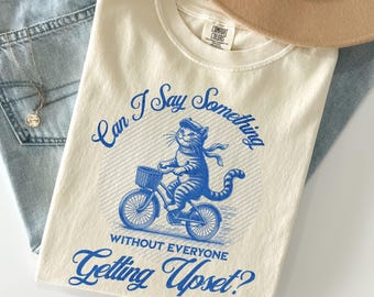 May include: Cream-colored t-shirt with a blue graphic of a cat riding a bicycle, with the text "Can I Say Something Without Everyone Getting Upset?" A tan hat and light blue jeans are in the background.