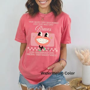 May include: A watermelon-colored t-shirt with the text "YOU CAN'T BUY HAPPINESS BUT YOU CAN BUY Braces AND HONESTLY IT'S KINDA THE SAME THING." The shirt features a cartoon graphic of a mouth with braces.