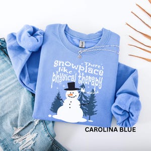 May include: Carolina blue sweatshirt with a snowman graphic and the text "Snowplace like physical therapy." The snowman has a black top hat and is surrounded by pine trees. A silver necklace is draped across the top.
