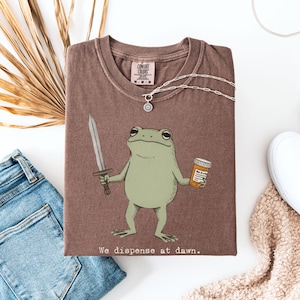 May include: A brown t-shirt featuring a cartoon frog holding a sword and a pill bottle. The text "We dispense at dawn" is printed at the bottom. A silver necklace and a pair of blue jeans are also visible.