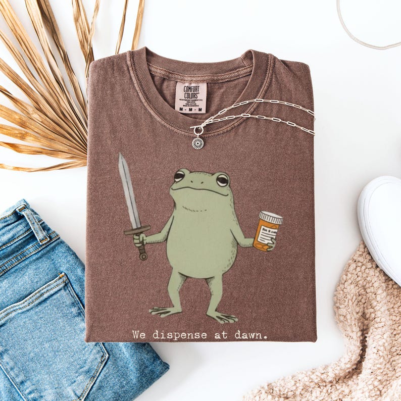 May include: A brown t-shirt featuring a cartoon frog holding a sword and a pill bottle. The text "We dispense at dawn" is printed at the bottom. A silver necklace and a pair of blue jeans are also visible.