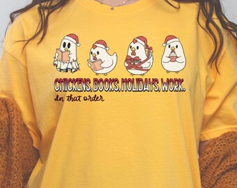 May include: Mustard yellow t-shirt featuring four cartoon chickens wearing Santa hats. Each chicken is engaged in a different activity: reading, gift-giving, and enjoying a beverage. The text "CHICKENS, BOOKS, HOLIDAYS, WORK. In that order" is printed below the chickens.