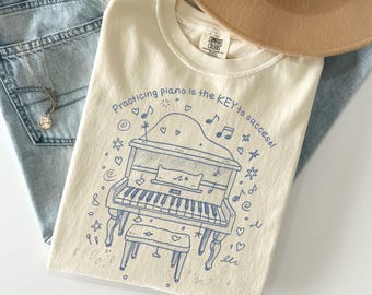 May include: Cream-colored t-shirt with a blue illustration of a piano and the text "Practicing piano is the KEY to success!". The shirt is paired with light blue jeans and a tan hat.