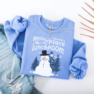 May include: A light blue sweatshirt with the text "snowplace like the lunchroom" and a snowman graphic. The snowman has a black top hat and is surrounded by pine trees. A silver necklace is draped across the sweatshirt.