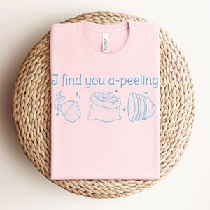 May include: A light pink t-shirt with the phrase "I find you a-peeling" in light blue script. The shirt features illustrations of potatoes and a potato sack. The shirt is folded on a woven, circular surface.