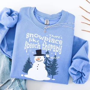 May include: A light blue sweatshirt with the text "snowplace like Speech therapy" and a snowman graphic. The snowman has a black top hat and is surrounded by pine trees. A silver necklace with a small pendant is on the sweatshirt.
