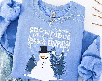 May include: A light blue sweatshirt with the text "snowplace like Speech therapy" and a snowman graphic. The snowman has a black top hat and is surrounded by pine trees. A silver necklace with a small pendant is on the sweatshirt.