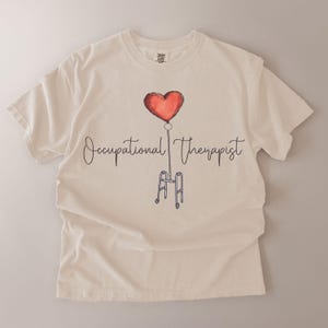 May include: Cream-colored t-shirt featuring a watercolor design. A red heart-shaped balloon is connected by a chain to a walker. The words "Occupational Therapist" are written in cursive below the balloon.