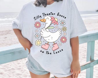 May include: Light blue t-shirt with a graphic of a white goose wearing pink boots and holding a flower. The text "Silly Theater Goose on the Loose" is printed above and below the goose, surrounded by colorful flowers.