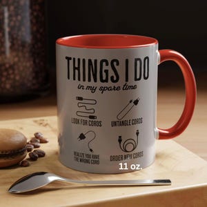 May include: A white ceramic mug with a red rim and handle. The mug has a black graphic that says "THINGS I DO in my spare time" and depicts a series of illustrations related to cords and cables. The text below the illustrations reads "LOOK FOR CORDS", "UNTANGLE CORDS", "REALIZE YOU HAVE THE WRONG CORD", "ORDER NEW CORDS", and "11 oz."