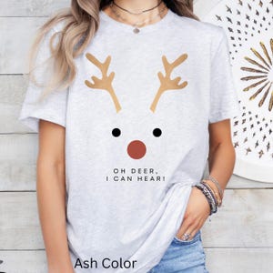 May include: Light gray t-shirt with a reindeer face design. The design includes brown antlers, black eyes, and a red nose. The text "OH DEER, I CAN HEAR!" is printed below the face. The shirt is labeled "Ash Color".