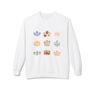 May include: White sweatshirt with a repeating pattern of colourful floral and fruit designs. The illustrations include tomatoes, wheat, and various flower arrangements in red, yellow, green, and blue. The sweatshirt has a crew neck and long sleeves.