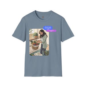 May include: A blue t-shirt featuring an illustration of a person in a kitchen with a basket of vegetables. Text bubbles read "Almost ready for the garden scene" and "Hope everyone tasted". The basket contains tomatoes, carrots, and other produce.