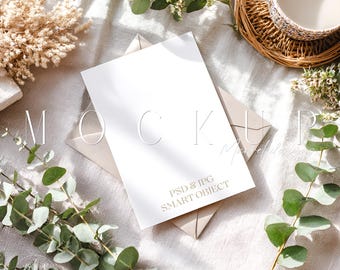 May include: A white card with the word "MOCKUP" and the text "PSD & JPG SMART OBJECT" is placed on a beige envelope. The scene is styled with eucalyptus, a cup of coffee, and dried flowers, creating a natural, aesthetic composition.