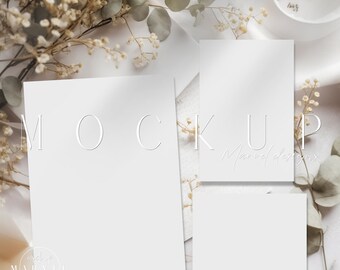 May include: A mockup featuring three blank white cards with a floral background. The cards are arranged in a vertical stack with the largest card at the bottom and the smallest card at the top. The background features white flowers and green leaves.