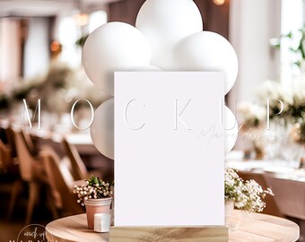 May include: White sign mockup with a wooden base, sitting on a wooden table with white balloons and greenery in the background. The sign says "MOCKUP" in a white font.