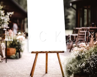 May include: A white sign on an easel with a wooden frame. The sign is blank and ready for text. The sign is in front of a wooden building and greenery.