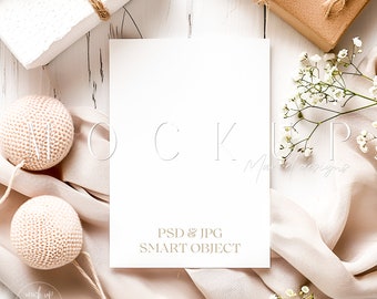 May include: A white card mockup with the text "PSD & JPG SMART OBJECT" on a white wooden background with a white gift box, a brown gift box, white flowers, and a beige fabric.