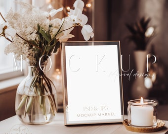 May include: A gold frame mockup with a white background, sitting on a wooden table with a vase of white flowers and a lit candle. The text "MOCKUP" is written in a thin, white font above the frame. The text "PSD & JPG MOCKUP MARVEL" is written in a thin, white font below the frame.
