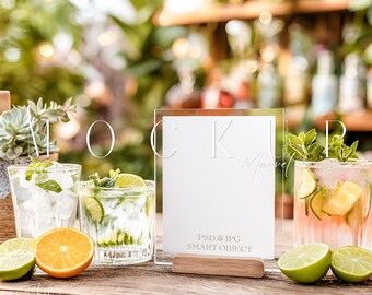 Può includere: Un cartello in acrilico trasparente con base in legno, che mostra il testo "MOCKUP Marvel" e "PSD & JPG SMART OBJECT". L'immagine presenta tre bicchieri di cocktail con lime e menta, e un'arancia e lime a fette su un tavolo di legno.