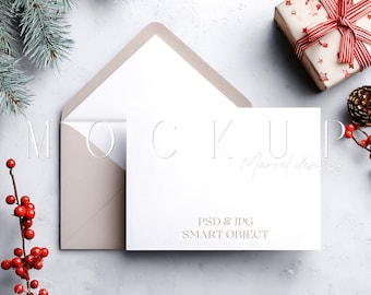 May include: A white card with the word "MOCKUP" embossed on it, inside a beige envelope. The card is on a light gray surface, with a wrapped gift, pine sprig, and red berries. Text on the card reads "PSD & JPG SMART OBJECT".
