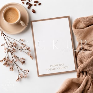Può includere: Un mockup di carta rettangolare con bordo marrone, una tazza di caffè beige su piattino abbinato, chicchi di caffè sparsi, un ramo di fiori secchi e un tessuto a maglia beige piegato. La carta riporta il testo MOCKUP, PSD & JPG SMART OBJECT.