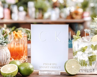 Può includere: Un cartello trasparente in acrilico con le lettere "CK" incise sulla superficie. Il cartello è esposto su un tavolo di legno accanto a un cocktail, un mojito e lime. Il testo "PSD & JPG SMART OBJECT" è stampato sul cartello.