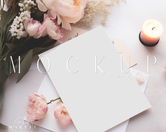 May include: A white card mockup with a blank white card on top of a white envelope. The card is surrounded by pink and white flowers and a lit candle. The text "MOCKUP" is written in white on the card.