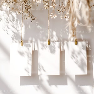 May include: Three blank white cards hanging from twine with gold clips against a white background. Dried flowers hang above the cards, casting shadows on the cards and background.