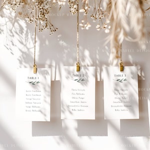 May include: Three white cards with gold clips hanging from twine. The cards are labeled "Table 1", "Table 2", and "Table 3" and list names for each table. The cards are hanging against a white background with dried flowers above them.