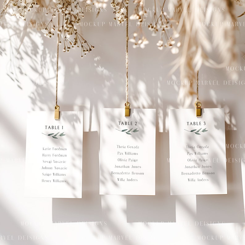 May include: Three white cards with gold clips hanging from twine. The cards are labeled "Table 1", "Table 2", and "Table 3" and list names for each table. The cards are hanging against a white background with dried flowers above them.