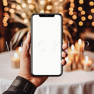 May include: A black smartphone with a blank white screen is held in a hand against a background of warm, blurry lights. The word "MOCKUP" is written in white text on the image.