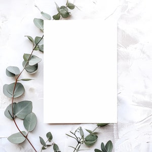 Puede incluir: Una maqueta de tarjeta blanca con una superficie blanca en blanco, rodeada de hojas de eucalipto verdes sobre un fondo blanco texturizado.