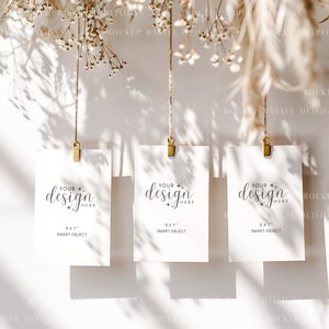 May include: Three white cards with gold clips hanging from twine against a white background. Each card has the text "Your design here" and "5x7" smart object.