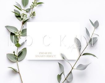 May include: A white card mockup with a simple, clean design. The card is surrounded by sprigs of green eucalyptus leaves on a white background. The text "MOCKUP" is printed in a light gray font on the card. The text "PSD & JPG SMART OBJECT" is printed in a smaller font below the word "MOCKUP".
