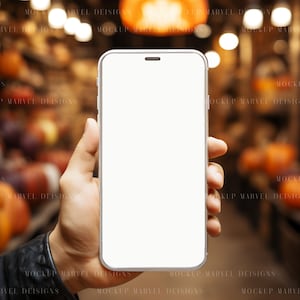 Op de afbeelding: Een hand die een witte smartphone vasthoudt met een leeg scherm. De achtergrond is wazig en toont een warme, uitnodigende sfeer met lichten en een rustieke omgeving.