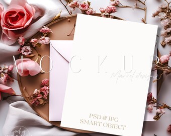 May include: A white card mockup with a pink envelope on a brown wooden tray. The tray is surrounded by pink flowers and a white fabric. The card has the text "PSD & JPG SMART OBJECT" printed on it.