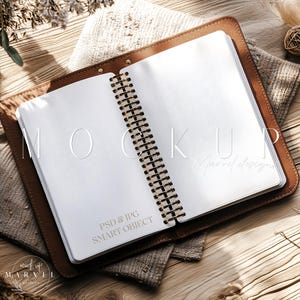 Puede incluir: Un cuaderno encuadernado en cuero marrón con espiral dorado, abierto en páginas blancas en blanco. El cuaderno está sobre una superficie texturizada beige. El texto "MOCKUP" y "Marvel Designs" es visible en las páginas.