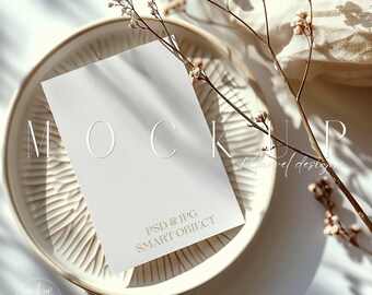 May include: A white card mockup with the text "MOCKUP" and "PSD & JPG SMART OBJECT" on a white plate with a sprig of dried white flowers.