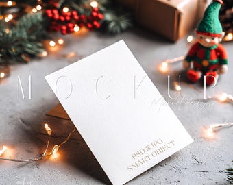 May include: A white card mockup with embossed text, angled on a wooden stand. The card is set against a festive backdrop with Christmas decorations, fairy lights, and a small elf figurine. The text "MOCK UP" is visible.