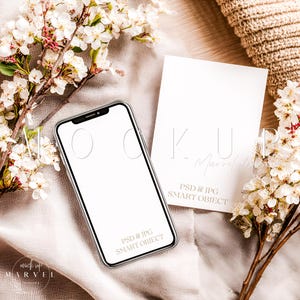 May include: A smartphone with a blank screen and a white card with the text "KUP Marvel designs" and "PSD & JPG SMART OBJECT" are displayed on a neutral fabric. White flowers and branches are arranged around the phone and card.