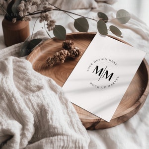May include: A white card with the text "Your Design Here" and the initials "M/M" in a circle with the text "Mock Up Marvel" below. The card is on a wooden tray with a white blanket and green leaves.