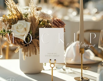 May include: A white card with the text "PSD & JPG SMART OBJECT" is displayed in a gold metal stand. The card is on a white tablecloth with a bouquet of dried flowers and a gold candle holder in the background.