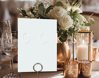 May include: A white card with a gold ring holder sits on a table with a bouquet of white roses and greenery. The card is blank and ready for a message. There are gold candle holders and votive candles on the table.