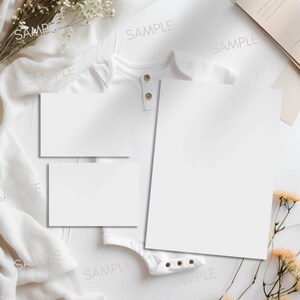Pode incluir: Dois cart&otilde;es brancos em branco, um maior que o outro, s&atilde;o colocados sobre uma superf&iacute;cie branca. Um macac&atilde;o branco para beb&ecirc; est&aacute; sobre a superf&iacute;cie, com os cart&otilde;es cobrindo-o parcialmente. O macac&atilde;o tem um fecho de bot&atilde;o e alguns bot&otilde;es pequenos na parte inferior. A superf&iacute;cie &eacute; decorada com tecido branco e flores secas.