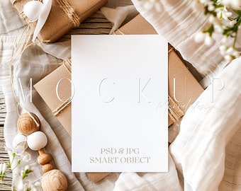 May include: A mockup scene featuring a white card with the word "MOCKUP" embossed on it, along with the text "PSD & JPG SMART OBJECT". The card is surrounded by wrapped brown boxes, white fabric, and wooden beads.