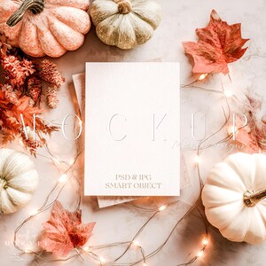 Könnte beinhalten: Eine Mockup-Szene mit einer leeren weißen Karte, auf der das Wort "MOCKUP" geprägt ist. Die Karte ist von kleinen Kürbissen, Herbstblättern und Lichterketten umgeben. Die Kürbisse sind in Weiß-, Pfirsich- und Goldtönen gehalten.