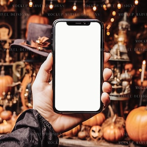 Op de afbeelding: Een hand die een zwarte smartphone met een wit scherm vasthoudt voor een Halloween-achtergrond met pompoenen, lampjes en decoraties.