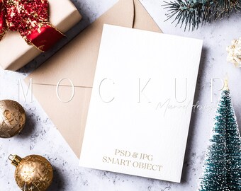 May include: A blank white card with the text "PSD & JPG SMART OBJECT" is propped against a tan envelope. The scene includes a wrapped gift with a red bow, gold ornaments, and a small Christmas tree, all on a textured surface.